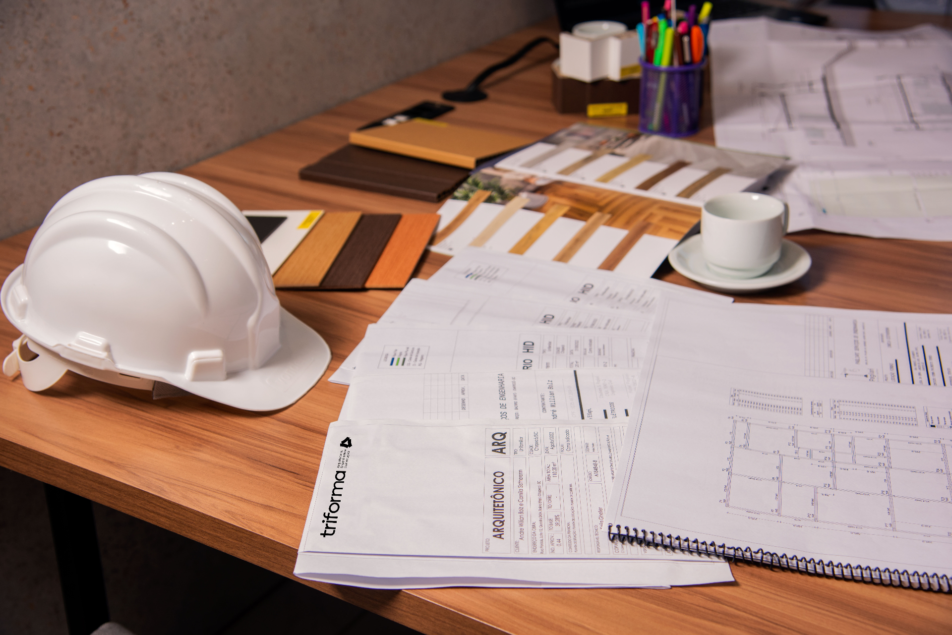 Pranchas técnicas, capacete e amostras de materiais sobre a mesa de projeto da Triforma